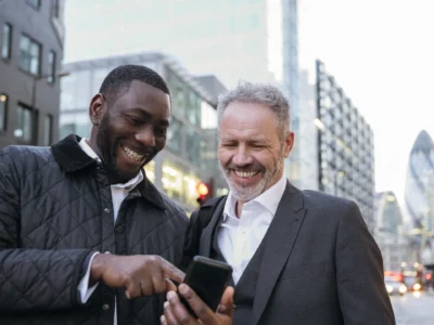 Two men in their 40s smiling together while looking at a phone on a London street, representing men's friendship and connection in midlife