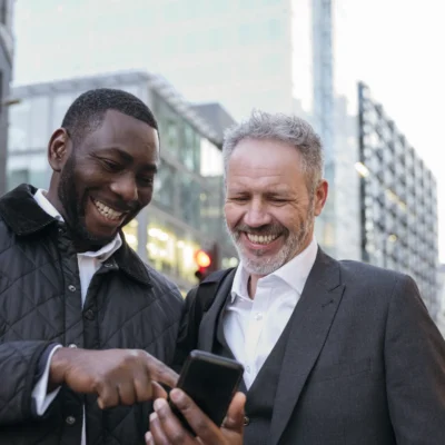 Two men in their 40s smiling together while looking at a phone on a London street, representing men's friendship and connection in midlife
