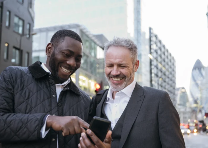 Two men in their 40s smiling together while looking at a phone on a London street, representing men's friendship and connection in midlife